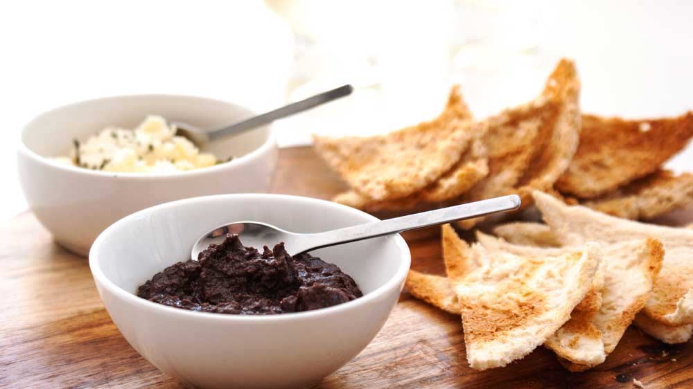 a white bowl of tapenade with toast behind