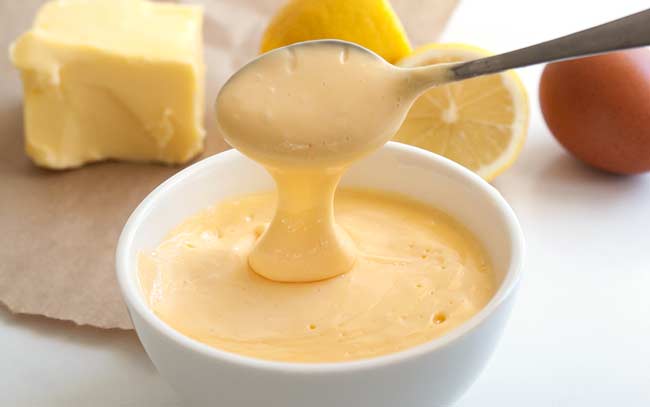 A spoon scooping up a dollop of easy Hollandaise sauce from a white bowl.