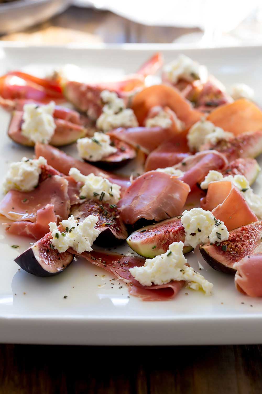 Large platter piled with figs, prosciutto, ricotta, and herbs on a wooden table