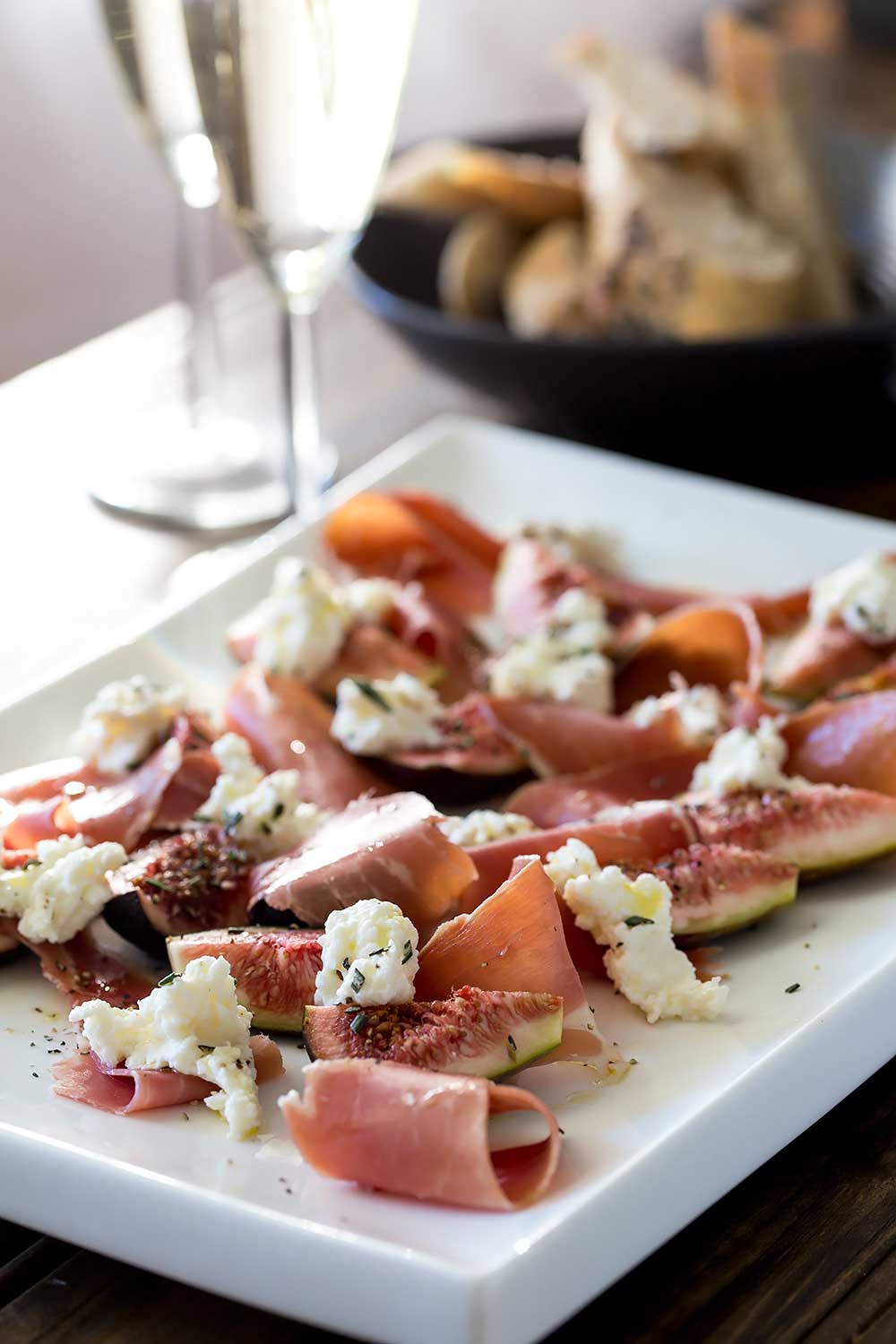 White plate of figs salad next to a bowl of bread with out of focus wine glasses behind.