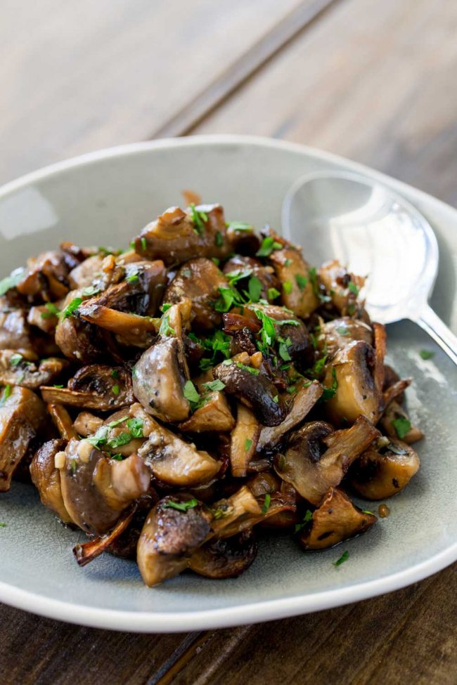 Grey marbled plate piled with roasted mushrooms, sprinkled with parsley. Silver serving spoon in the background