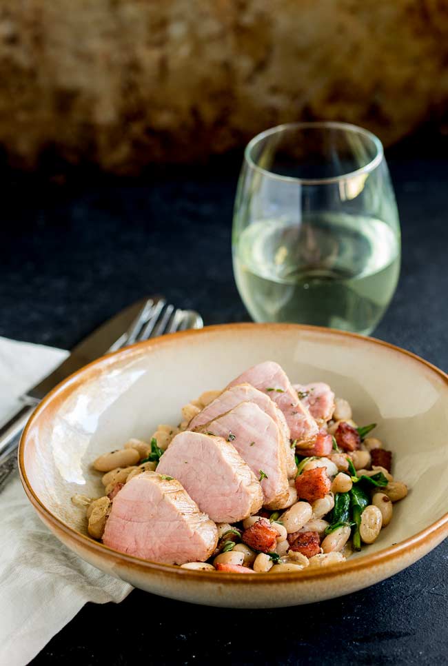 Full table view of a earthenware bowl, filled with white beans, crispy bacon and wilted spinach. Wine glass in the background.