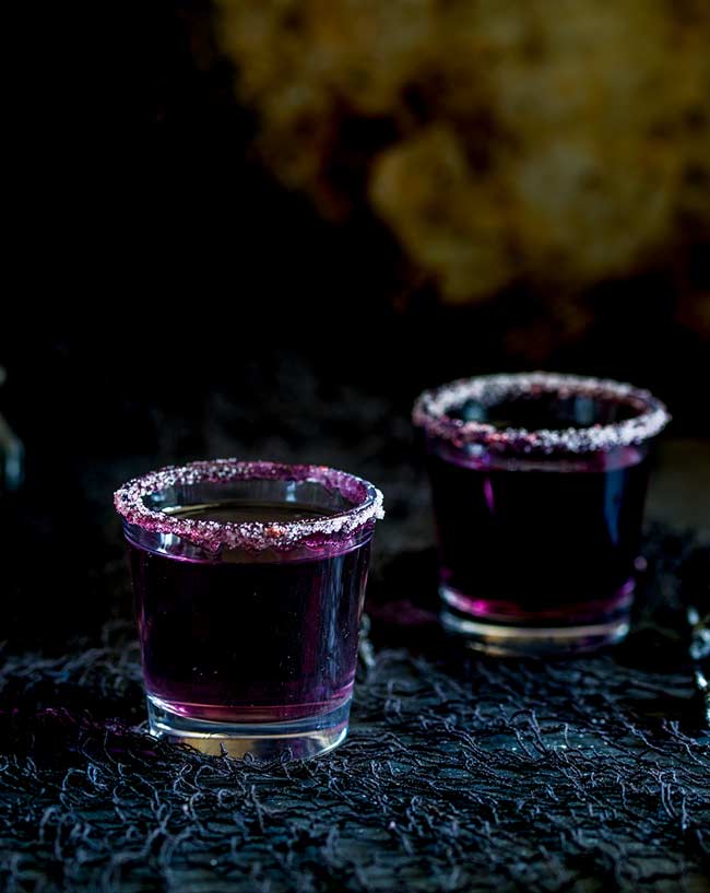 Two shot glasses, with sugar rim and a purple cocktail in them, on a black table.