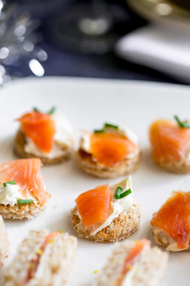 Round toasts spread with cream cheese and topped with smoked salmon and chives. On a white plate.