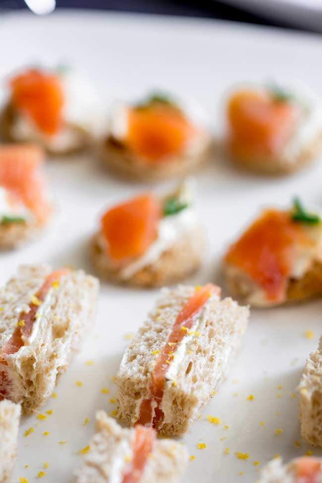 Brown bread with lemon cream cheese and and smoked salmon finger sandwiches on a large white plate. Pasta bites in the background.