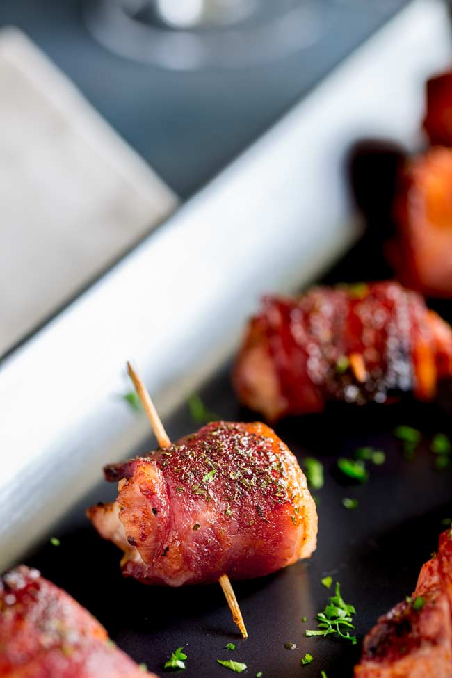 Close up on a chicken bite. Crispy bacon wrapped chicken with a cocktail stick, sat on a black plate and garnished with parsley.