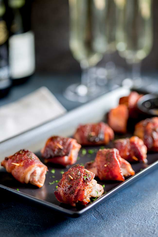 Retangular black plate with 9 Brown Sugar Bacon Wrapped Chicken Bites on it, wine bottle and wine glasses in the background all on a dark blue table.