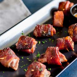 Sure picture of Brown Sugar Bacon Wrapped Chicken Bites on a black rectangular plate with a white napkin in the background.