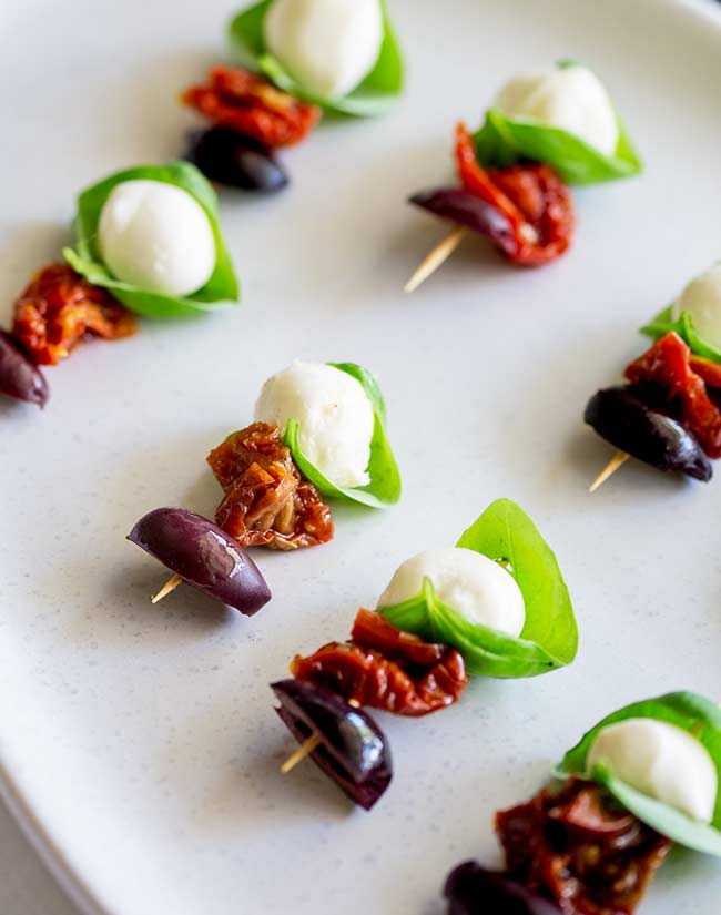 Mini mozzarella wrapped in basil leaf skewered with a sundried tomato and olive half, on a rustic white platter.