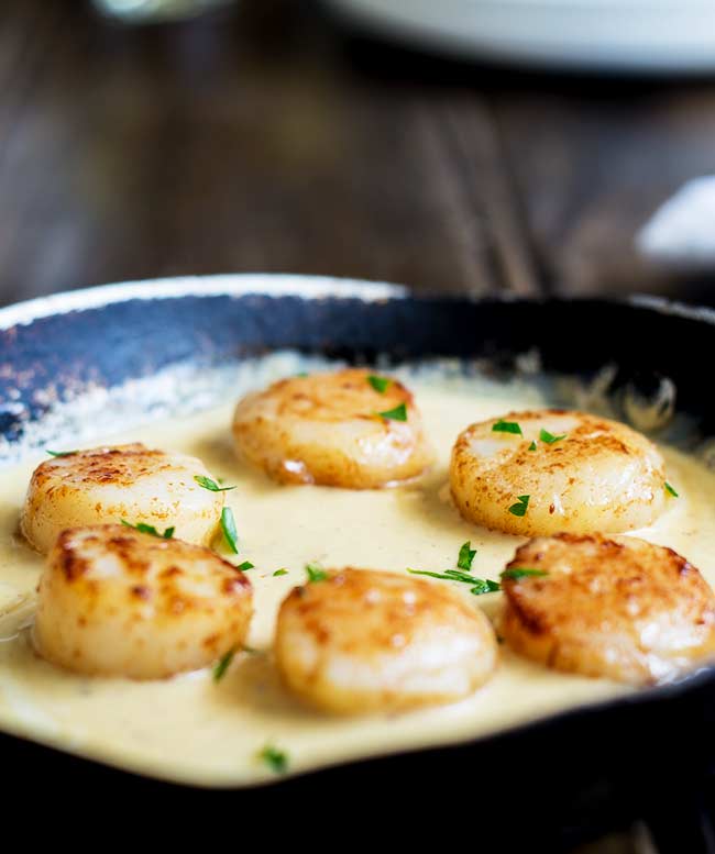 Close up of seared scallops in a small cast iron pan with a saffron cream sauce over them and parsley on top.