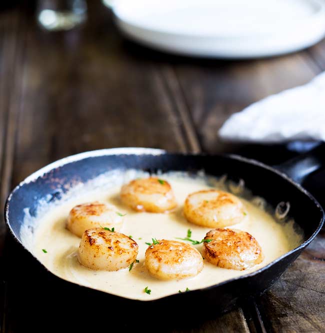 Small fry pan with seared scallops sat in a saffron cream sauce with white plates and napkins in the background.