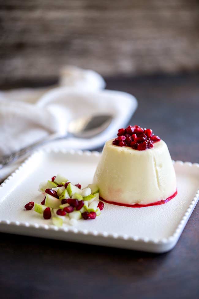 Square white frilly edged plate, with one pear and pomegranate panna cotta on it with a pear and pomegranate relish piled at the side and pomegranate arils on top.