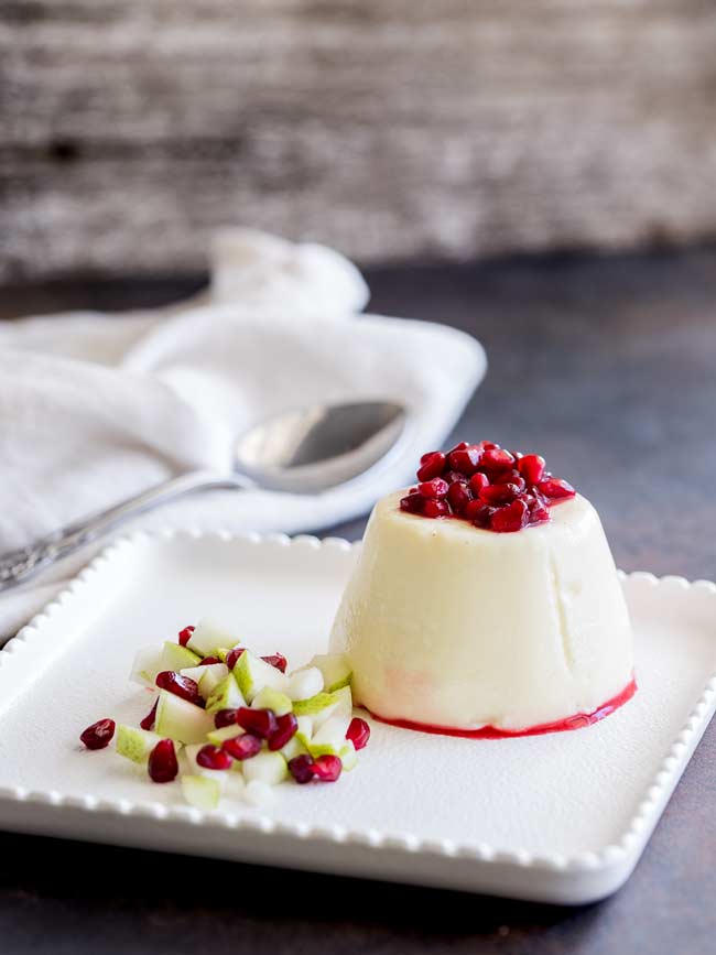 A Pear and Pomegranate Panna Cotta sat on a white plate with a crisp white napkin behind and silver spoon. Pomegranate sauce is dribbling down the pear panna cotta.