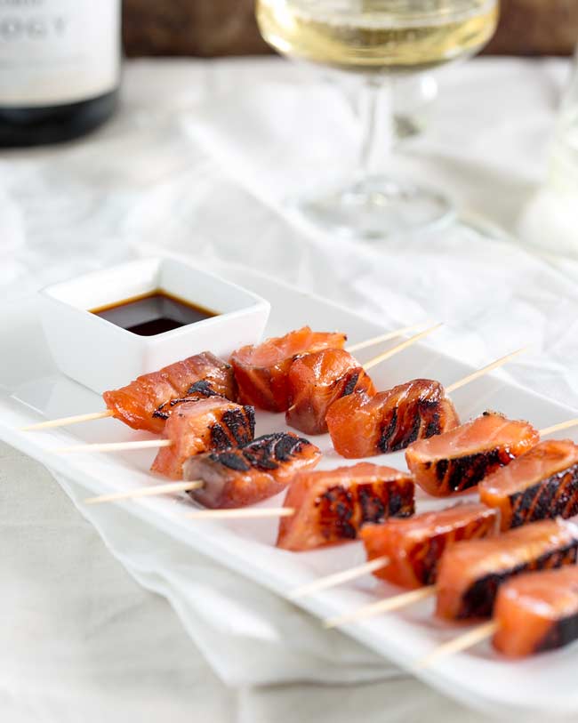 Long white platter, with cocktail stick skewered salmon bites, on a white table with champagne in the background.