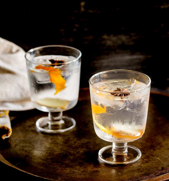 Two stemmed glasses with gin and tonic in them, garnished with star anise and orange zest. On an old metal tray with dark background