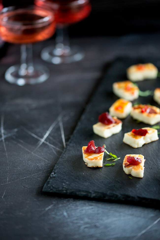 A slate platter, with small chunks of grilled halloumi cheese dressed with red cranberry sauce, garnished with mint. In the background 2 champagne glasses.