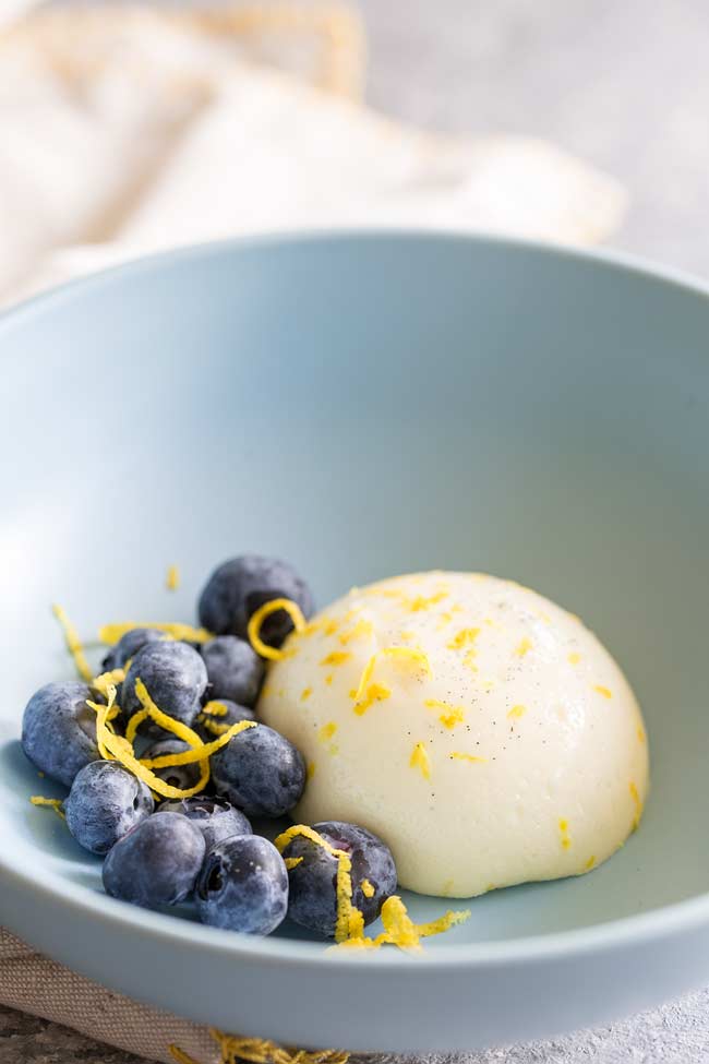 An unmoulded lemon panna cotta, garnished with blueberries and lemon zest in a shallow blue bowl.