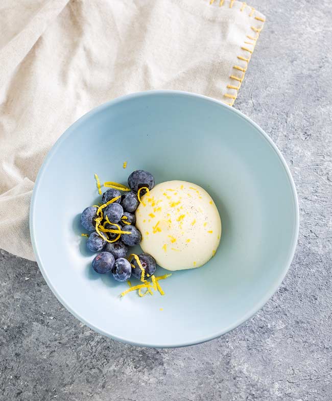 Overhead view, lemon panna cotta in a pale blue bowl sat on a linen napkin.