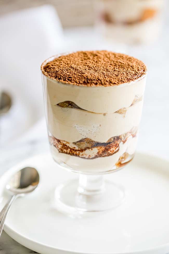 Individual tiramisu in a glass sat on a white plate, with silver spoon next to it.