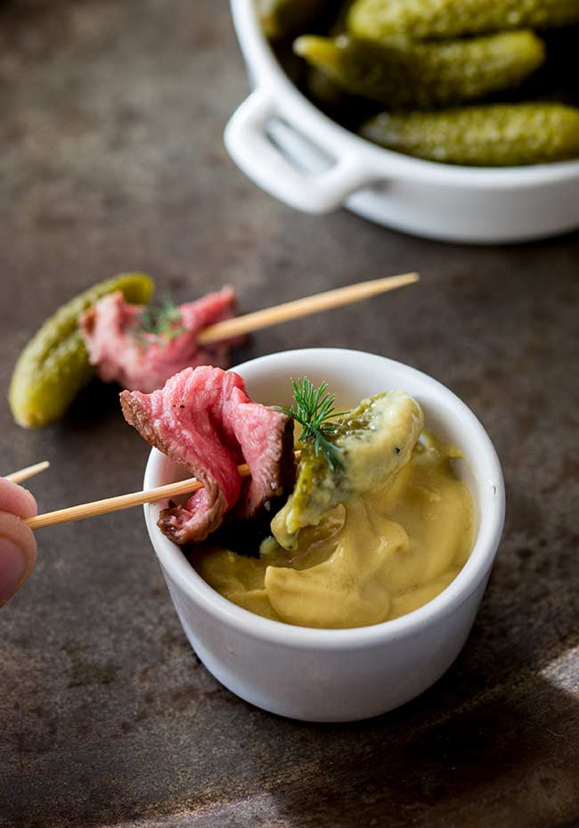 Dill pickle beef skewer being dipped into a small pot of sweet mustard.