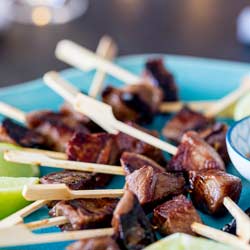 SQ picture showing the lamb skewers on a turquoise plate, garnished with lime with fairy lights in the background.
