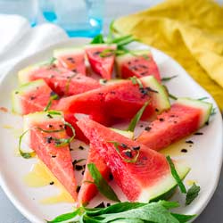 A SQ overhead shot of salted watermelon on a white plate, with a yellow napkin and blue glasses