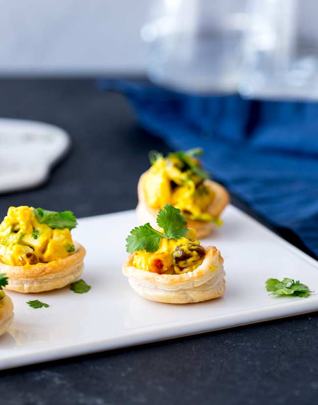 White platter on a black table with 4 pastry cups filled with yellow curried chicken and garnished with cilantro