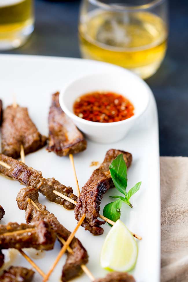 Thai beef on a wooden skewer on a white platter with a red chili dipping sauce in a white bowl