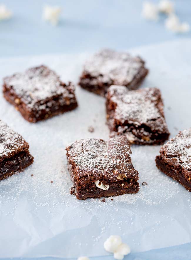 6 popcorn brownie squares on a sheet of parchment on a blue table.