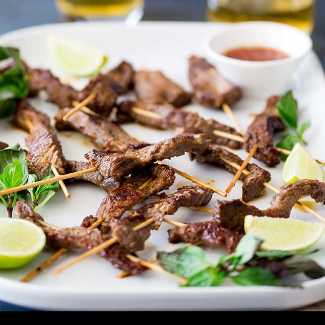 Thai beef skewers on a white platter, garnished with thai basil and lime segments