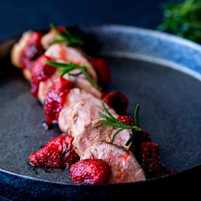 SQ image showing 10 slices of pork tenderloin and arranged around the slices are roasted strawberries and rosemary leaves