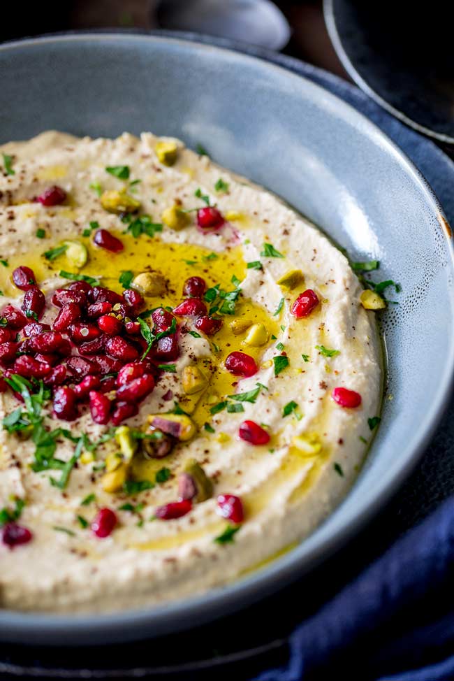 hummus in a flat blue bowl with pomegranate, pistachio and parsley scattered over the top
