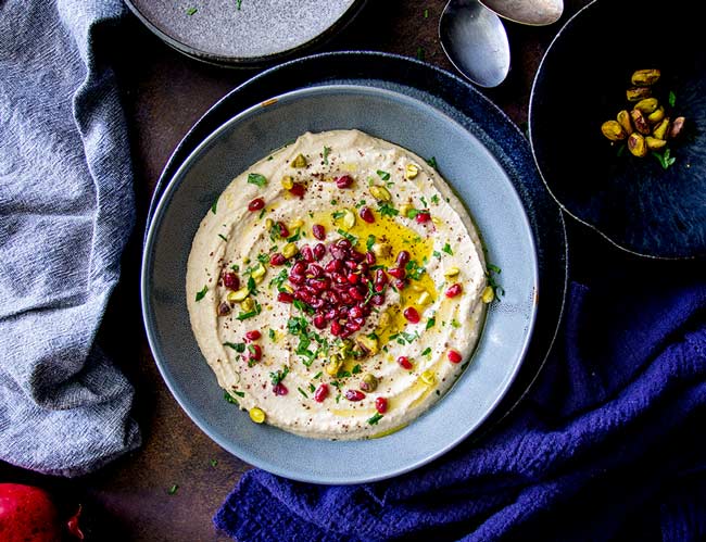 overhead head view of hummus in a blue bowl on a dark table with spoons and rustic lines around the edge.