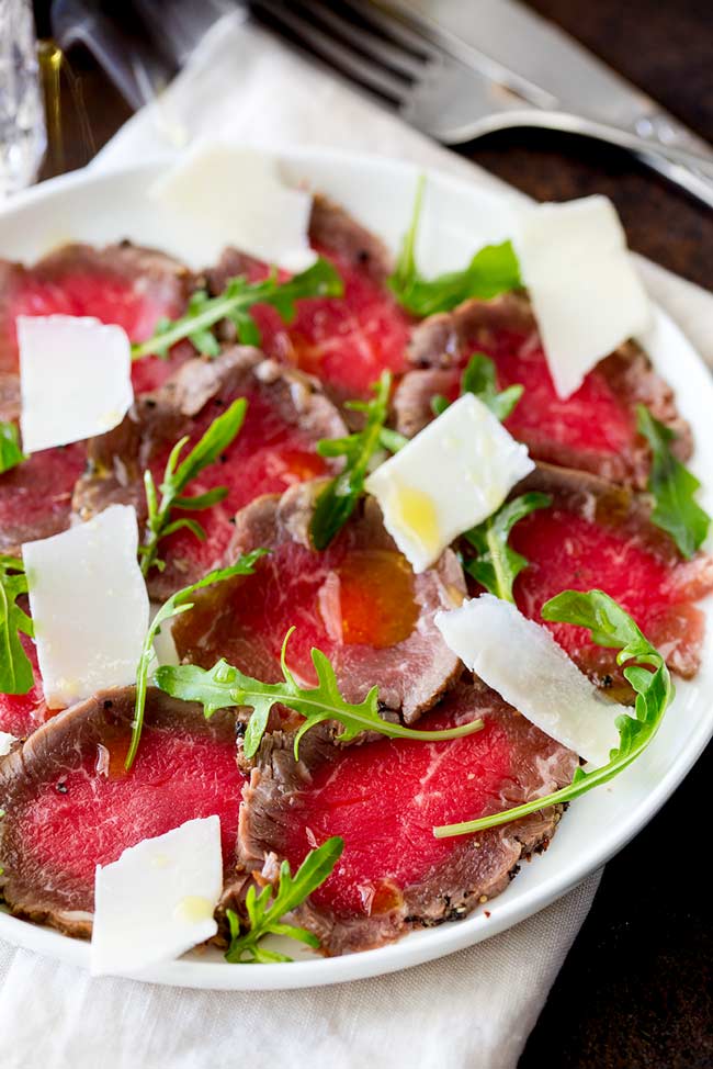Close up on the thin slices of beef carpaccio garnished with baby arugula and shaved parmesan.