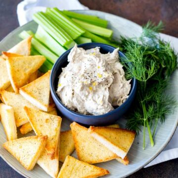 table view, grey plate on a brown table with a blue bowl of smoked fish pate on it and vegetable batons and toast points arranged around the plate
