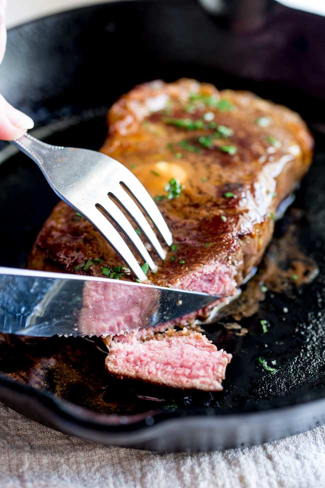 a knife and fork cutting a strip from a ribeye steak in a cast iron pan