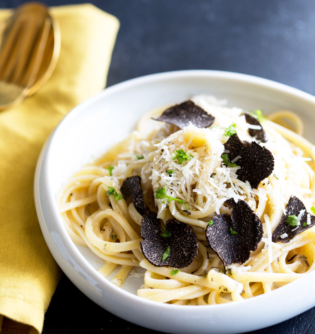 overhead of a shallow white bowl of black truffle pasta, garnished with parmesan and shaved black truffle