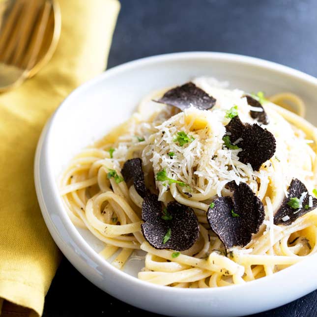 Square image of a flat shallow bowl of black truffle pasta on a black table with a yellow napkin
