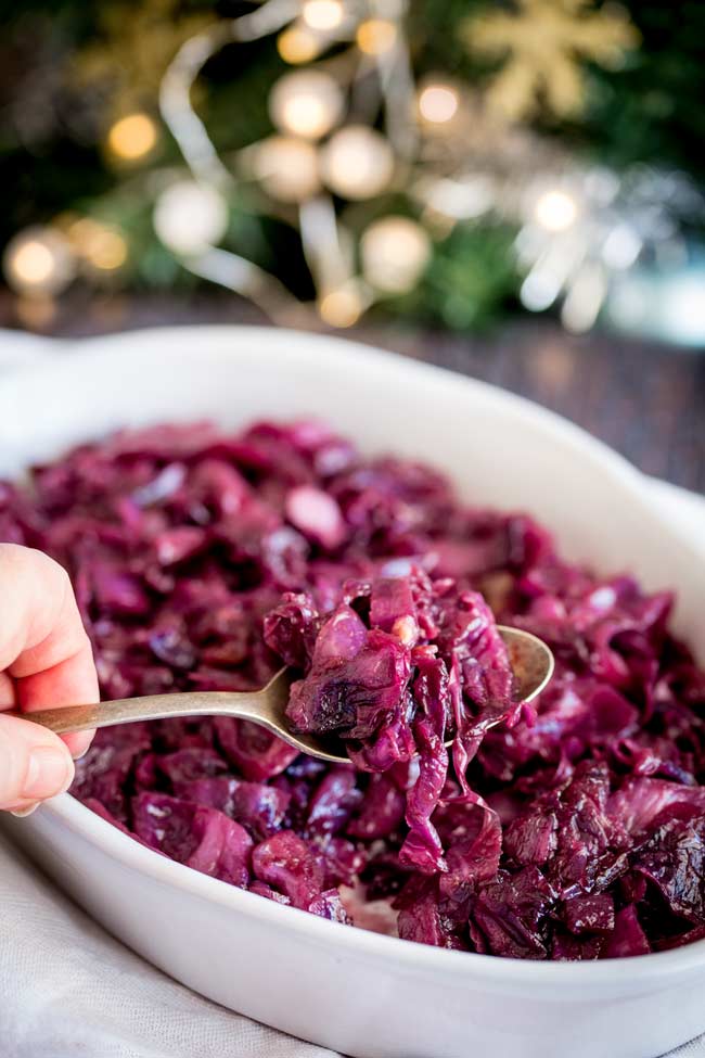 a hand lifting a spoonful of braised red cabbage from a white serving dish