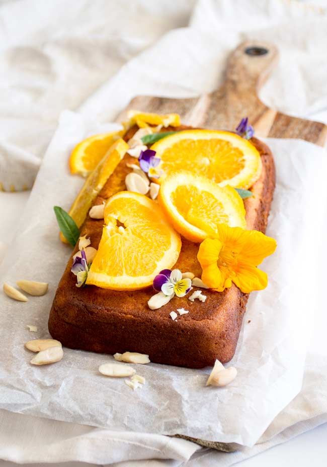 a long thin cake on a wooden board garnished with slices of orange, almonds and purple flowers