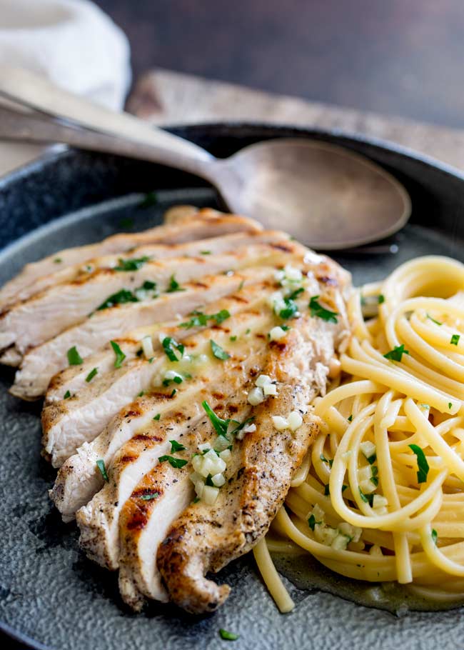 looking down on a sliced chicken breast with garlic butter next to a pile of garlic butter pasta on a metal place with a worn spoon