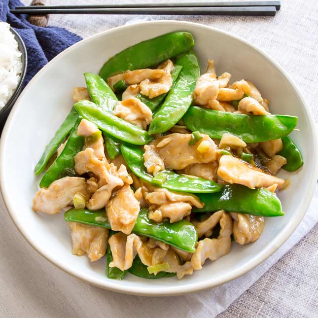 takeout style chicken and snow pea stir fry in a white bowl with a bowl of rice and chopsticks
