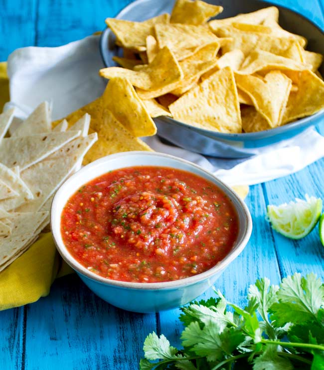 bright blue table with a bowl of salsa, bowl of chips, lime and fresh cilantro, on it.