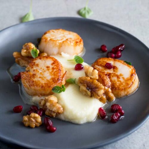 square picture of 3 seared scallops with cauliflower puree on a grey plate garnished with walnuts, pomegranate and herbs
