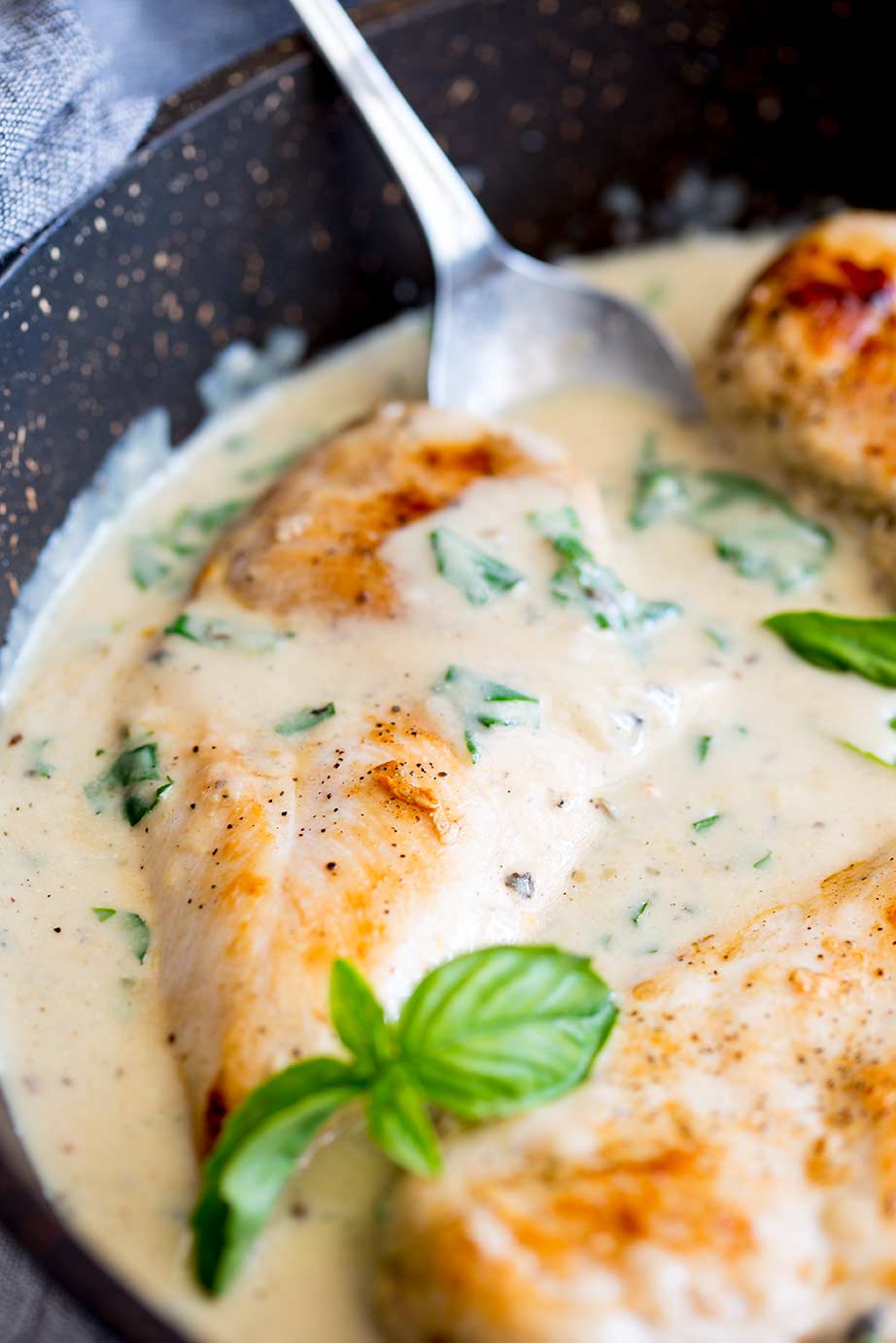 a chicken breast in a pan of creamy sauce with a silver spoon in the background