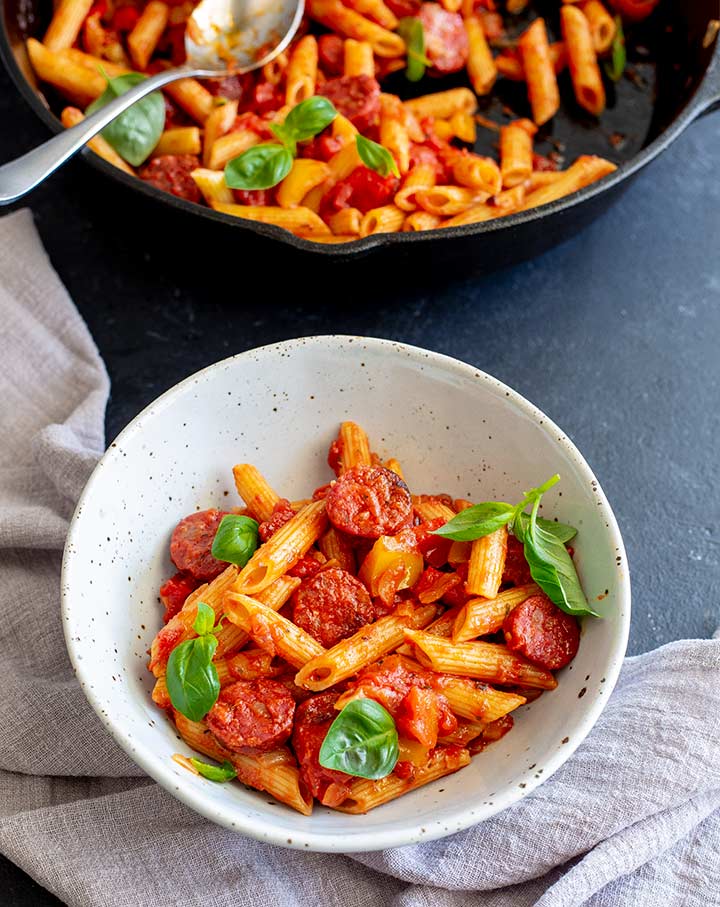 a rustic bowl of chorizo pasta on a black table with a pan in the background