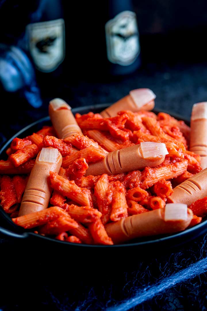 A black bowl of red pasta with frankfurter sausage fingers with potion bottles in the background