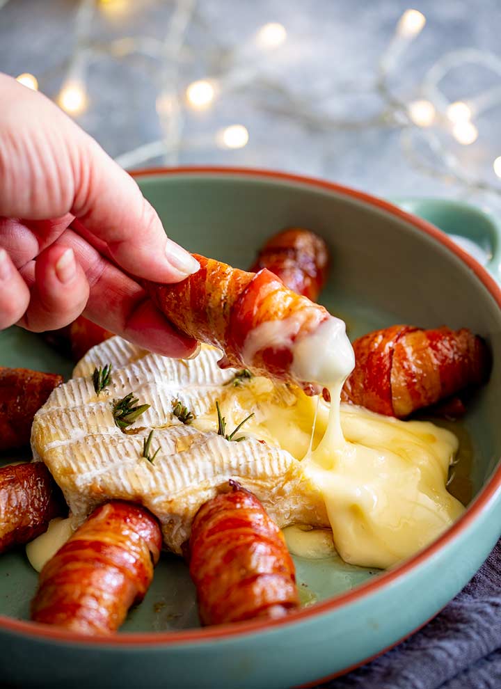 a hand dripping a bacon wrapped sausage into molten brie