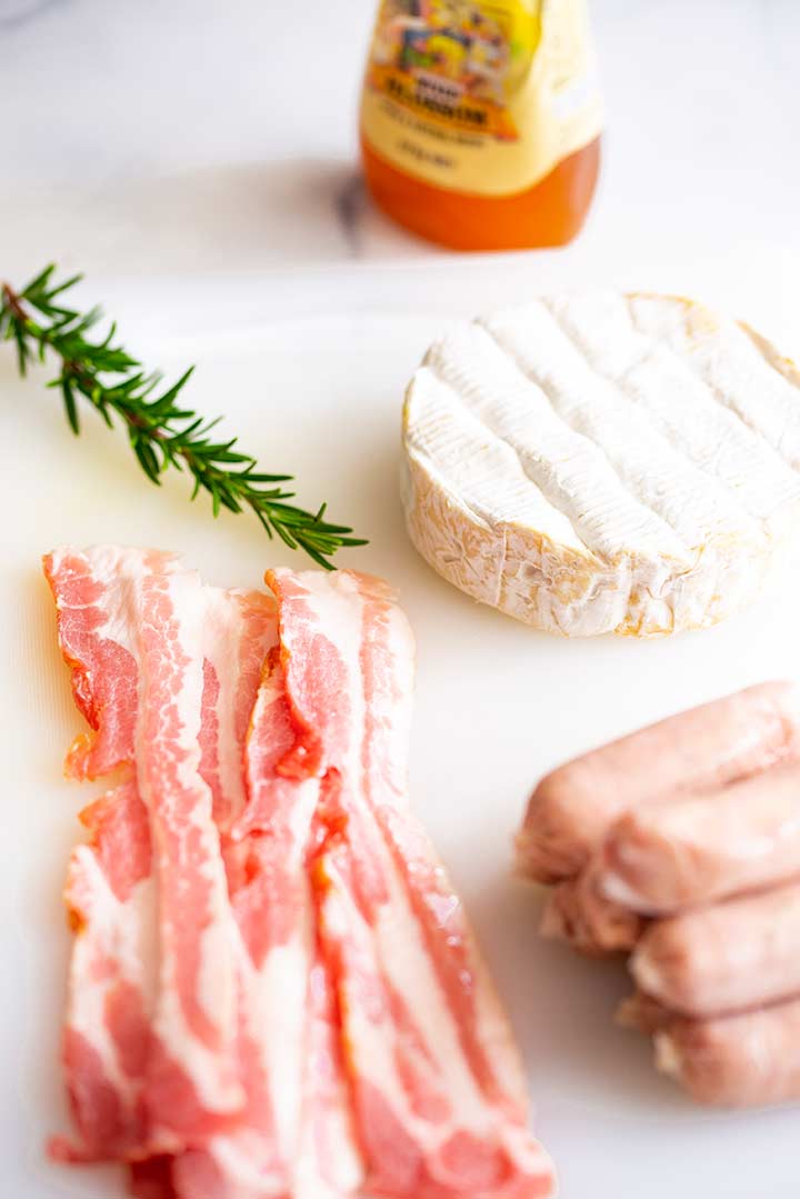 sausages, bacon, brie, rosemary and honey sitting on a cutting board on a marble table