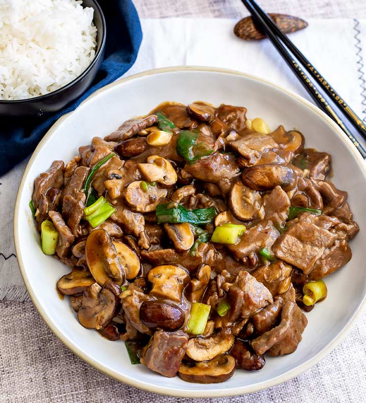 overhead of a white bowl of Chinese beef and mushrooms with rice and chop sticks behind